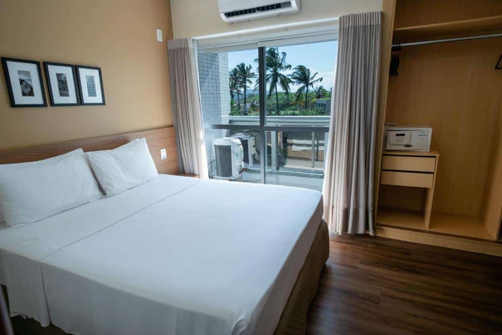 Quarto no Nobile Suites Diamond com uma cama de casal, armário com cofre e uma janela com cortinas que leva até uma varanda com vista para a cidade, para representar hotéis em Vitória.