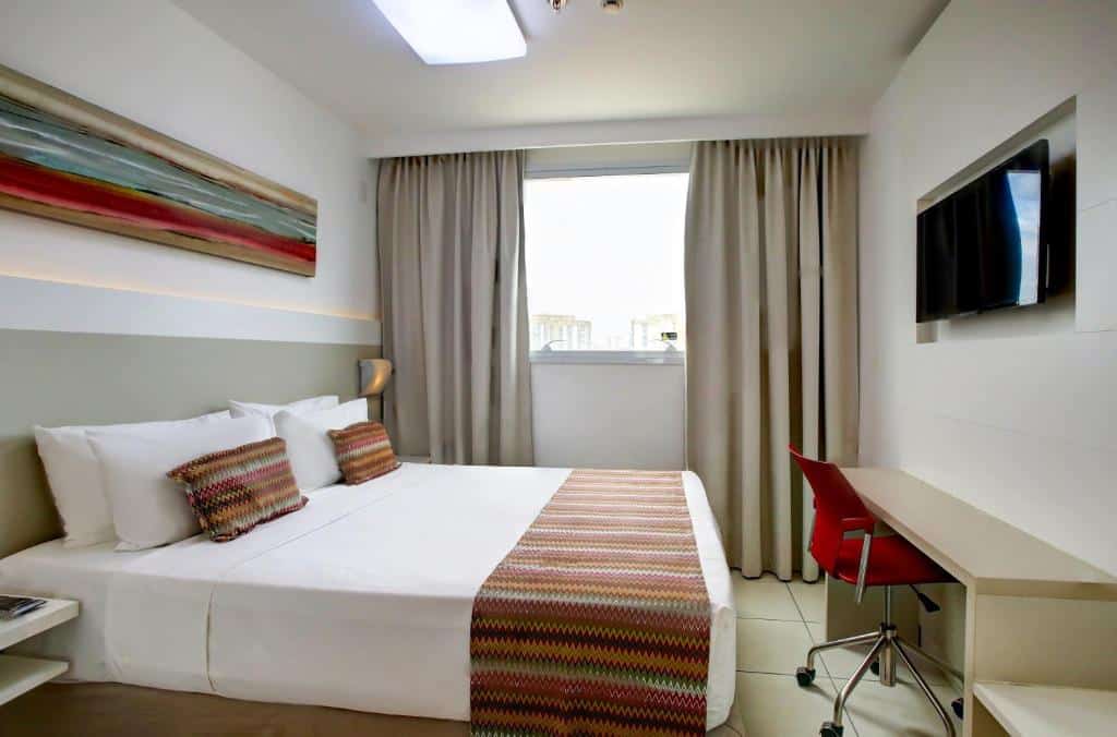 Quarto no Easy Vitória Reta da Penha by Atlantica com uma cama de casal, mesa com cadeira, televisão e janela com cortinas e vista da cidade, para representar hotéis em Vitória.