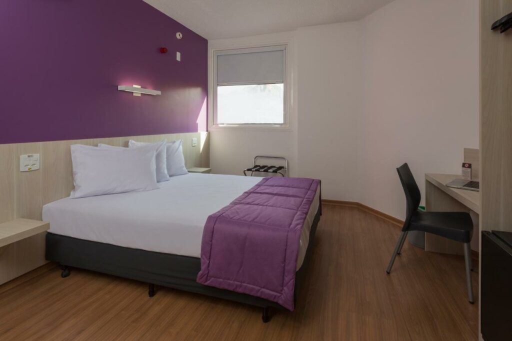 Quarto no Go Inn Vitória com uma cama de casal, mesa com cadeira e uma pequena janela. Uma parte da parede e a colcha são roxas, para representar hotéis em Vitória.