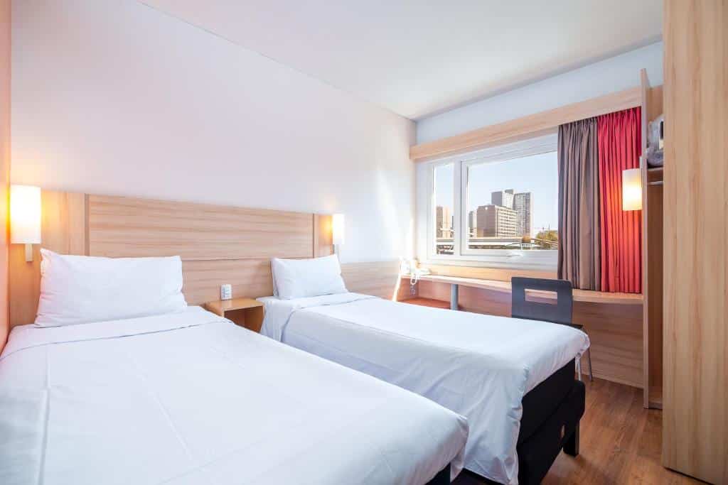Quarto do ibis Santiago Estacion Central com uma janela ampla com cortinas, uma bancada de trabalho com uma cadeira, duas camas de solteiro, um chão de madeira, para representar hotéis Ibis em Santiago do Chile