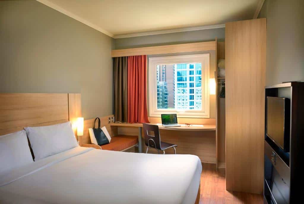 Quarto do ibis Santiago Las Condes Manquehue com uma cama de casal, uma janela com cortinas, uma mesa de escritório com uma cadeira, um pequeno sofá, uma televisão e um pequeno armário 
