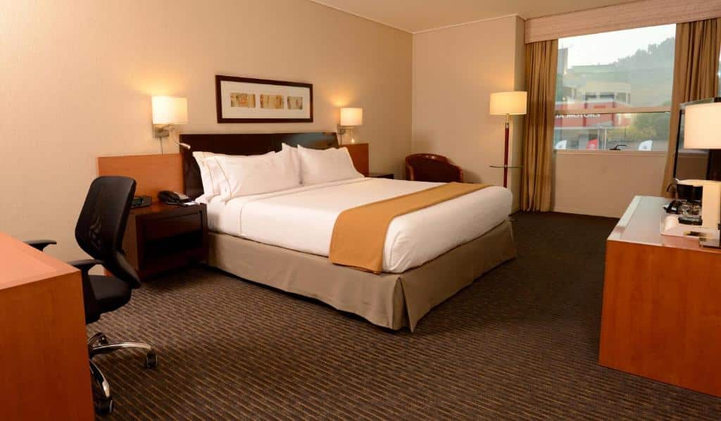 Quarto do Holiday Inn Express Santiago Las Condes, an IHG Hotel com cama de casal do lado esquerdo, duas cômodas ao lado da cama e em frente a cama uma mesa com TV.