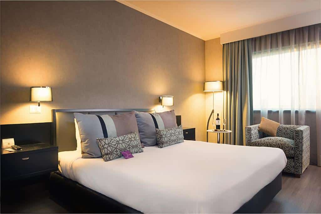 Quarto Mercure Santiago Centro com cama de casal no lado esquerdo, duas cômodas ao lado da cama e uma poltrona do lado direito. 