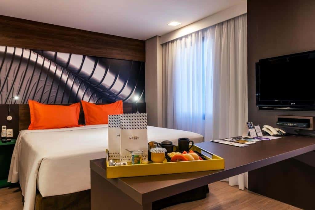 Quarto do hotel com uma cama de casal, travesseiros laranjas, tv na parede, quadro atrás da cama e uma bandeja amarela com comidas.