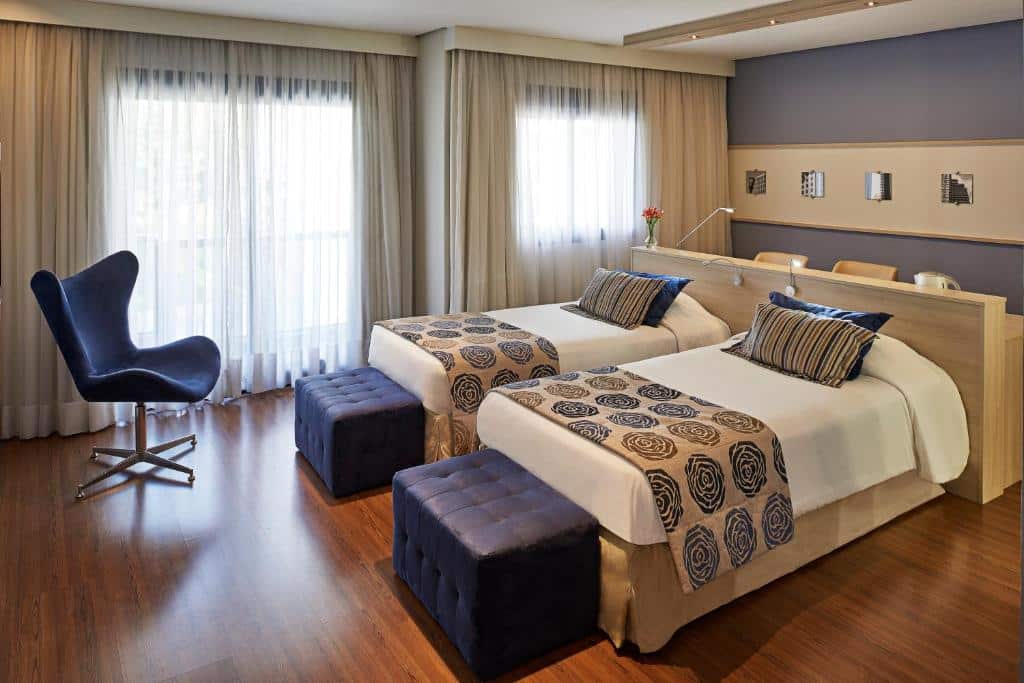 Quarto com duas camas de solteiro, dois puff no pé da cama, uma poltrona, piso de madeira e duas janelas com cortinas.