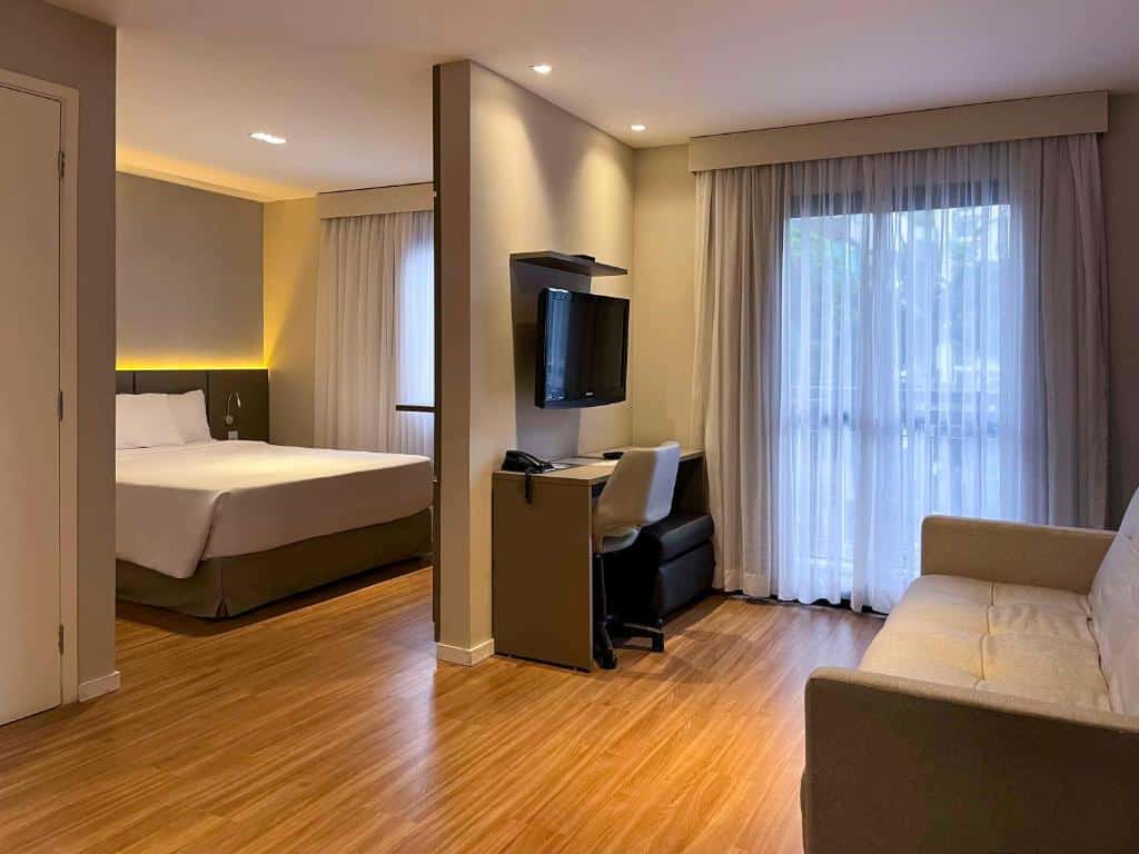 Quarto do hotel com uma cama ao fundo e uma porta separando o quarto para dormir com a sala de estar com uma tv na parede, mesa com cadeira, um sofá e uma janela de vidro com cortina.