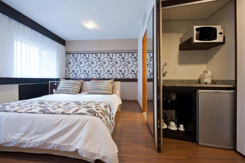 Quarto do hotel com uma cama de casal, uma janela com cortina, piso de madeira e ao lado uma parte com micro-ondas, frigobar e algumas canecas.