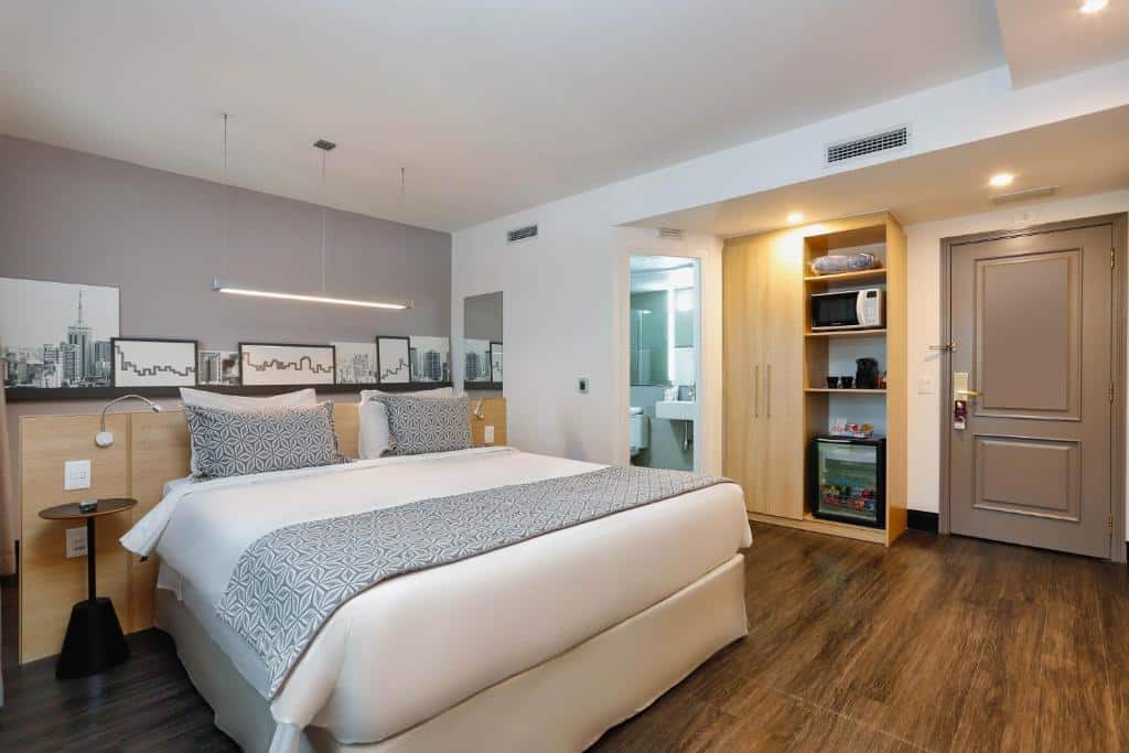 Quarto com uma cama de casal com piso de madeira, alguns quadros na parede, porta que da para o banheiro, um armário de madeira e uma porta marrom.