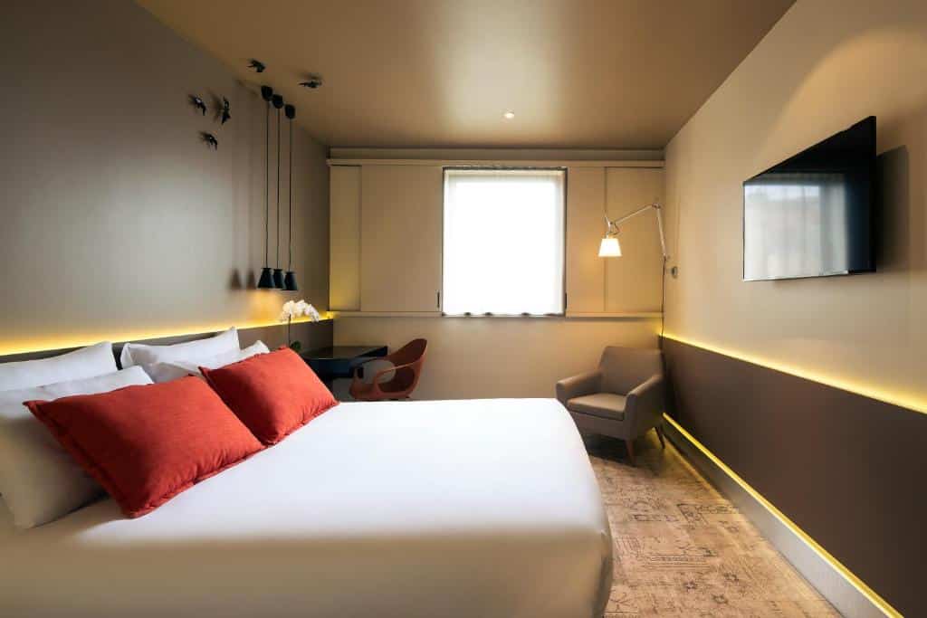 Quarto do Mercure Porto Centro Santa Catarina com cama de casal do lado esquerdo da imagem, em frente a cama uma TV presa na parede, do lado esquerdo da cama uma mesa de trabalho com cadeira e um poltrona a frente. Representa hotéis Mercure no Porto.