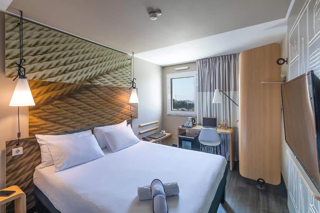Quarto do Hotel ibis Porto Gaia com cama de casal do lado esquerdo da imagem com duas cômodas em cada lado da cama, em frente a cama TV presa na parede e do lado esquerdo da cama mesa de trabalho com cadeira. Representa hotéis Mercure no Porto.