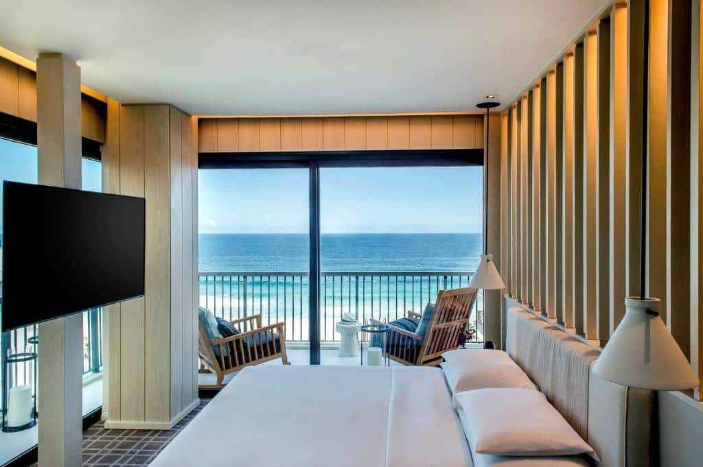 Quarto de hotel no Grand Hyatt com uma cama, duas cadeiras em uma varanda, janelas grandes e vista para o oceano sob um céu claro, para representar hotéis na Barra da Tijuca no RJ.