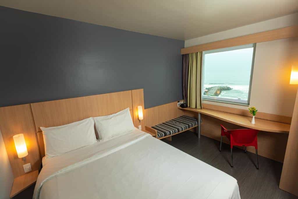 Um quarto moderno no ibis Barra da Tijuca com uma cama de casal, escrivaninha, cadeira vermelha, banco e janela com vista para o mar, para representar hotéis na Barra da Tijuca no RJ.