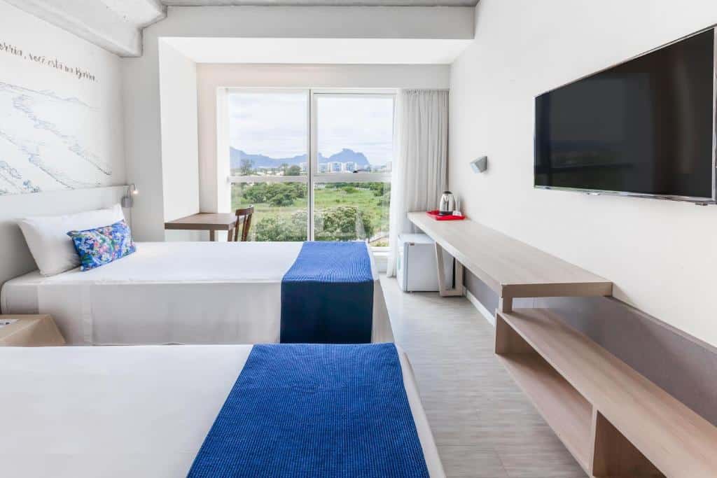 Quarto do Ramada Encore by Wyndham Ribalta com duas camas, detalhes em azul, uma TV na parede, uma escrivaninha e uma janela com vista para a vegetação e as montanhas, para representar hotéis na Barra da Tijuca no RJ.