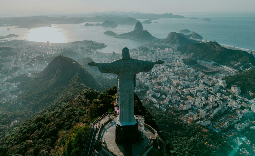 Estátua do Cristo Redentor vista de costas no Rio de Janeiro. Ao fundo fica o mar e alguns montes, e ao lado direito é possível ver casas e prédios na cidade.