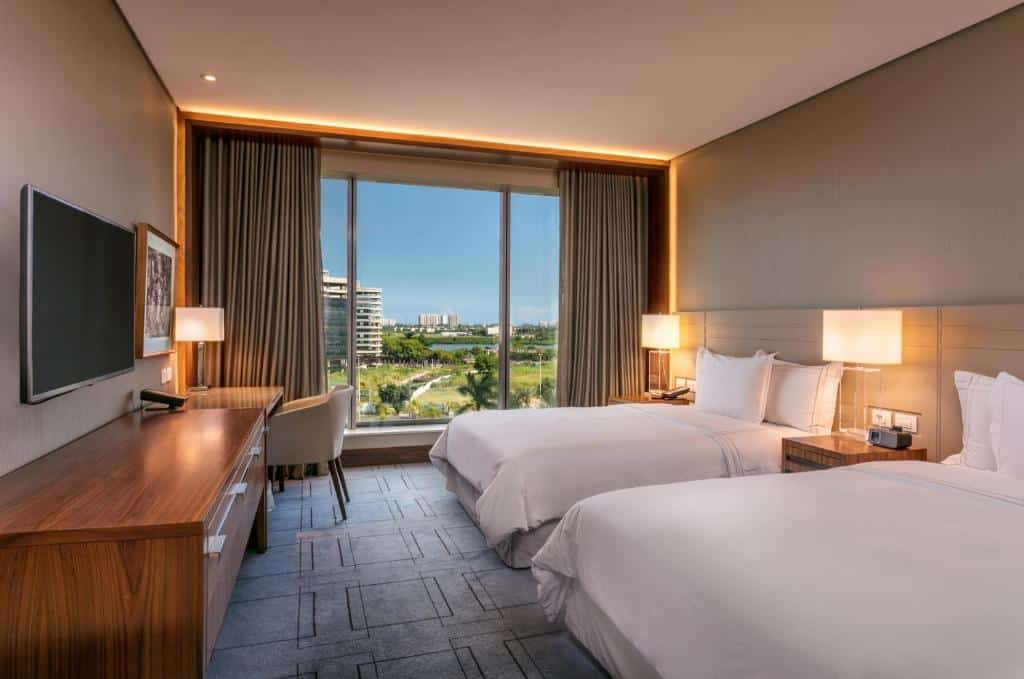 Quarto do Hilton Barra com duas camas de casal, uma escrivaninha e cadeira, uma TV de tela plana e uma janela grande com vista para edifícios e vegetação, para representar hotéis na Barra da Tijuca no RJ.