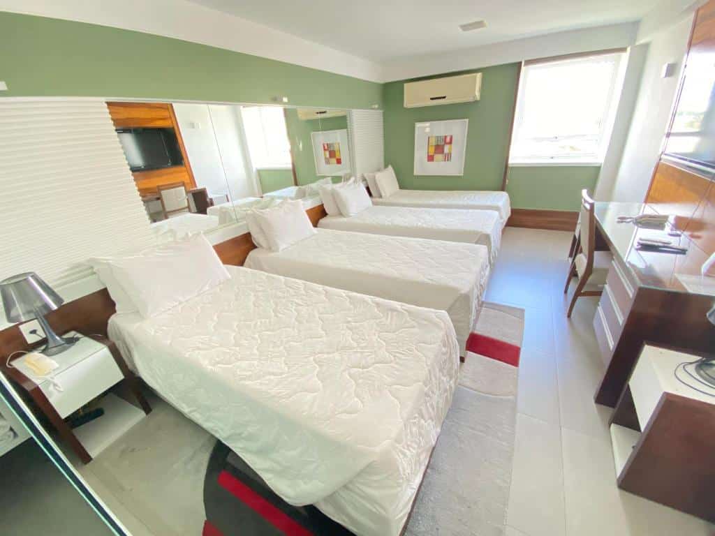 Quarto do TRYP by Wyndham Barra no Parque Olímpico com três camas de solteiro, uma TV na parede, uma escrivaninha, um abajur e uma janela que permite a entrada de luz natural, para representar hotéis na Barra da Tijuca no RJ.
