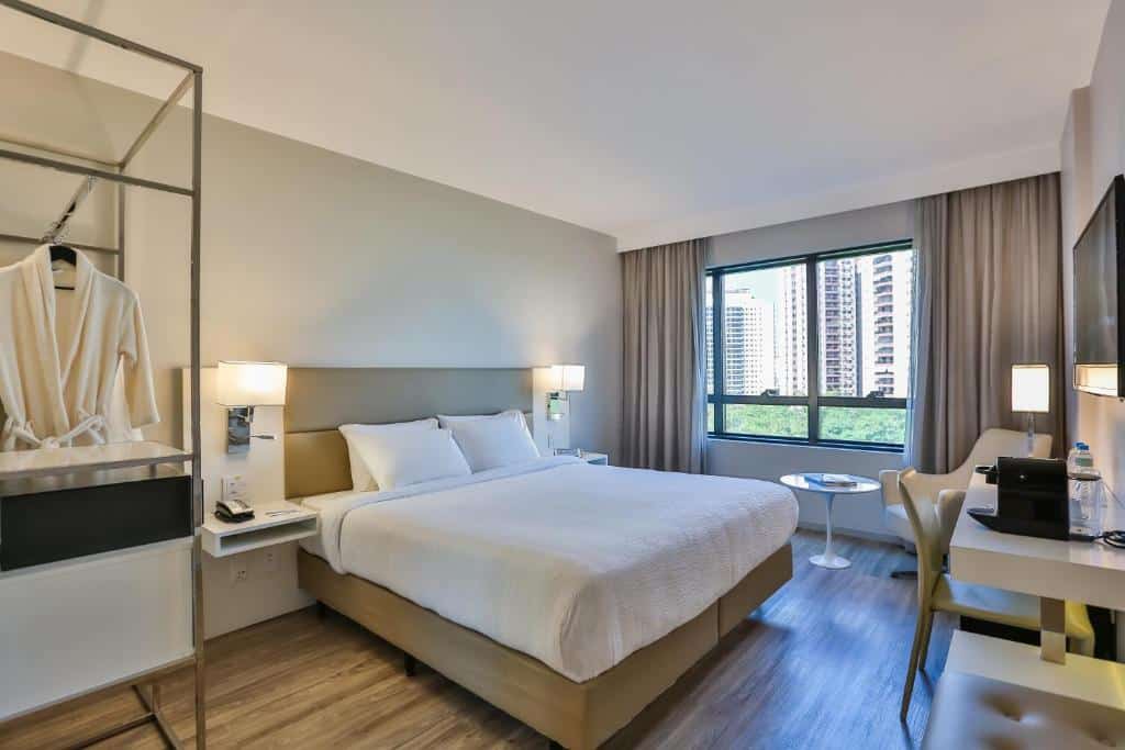 Quarto moderno no Radisson Barra com uma cama king-size, escrivaninha, cadeira e vista da janela para os prédios da cidade e a vegetação, para representar hotéis na Barra da Tijuca no RJ.