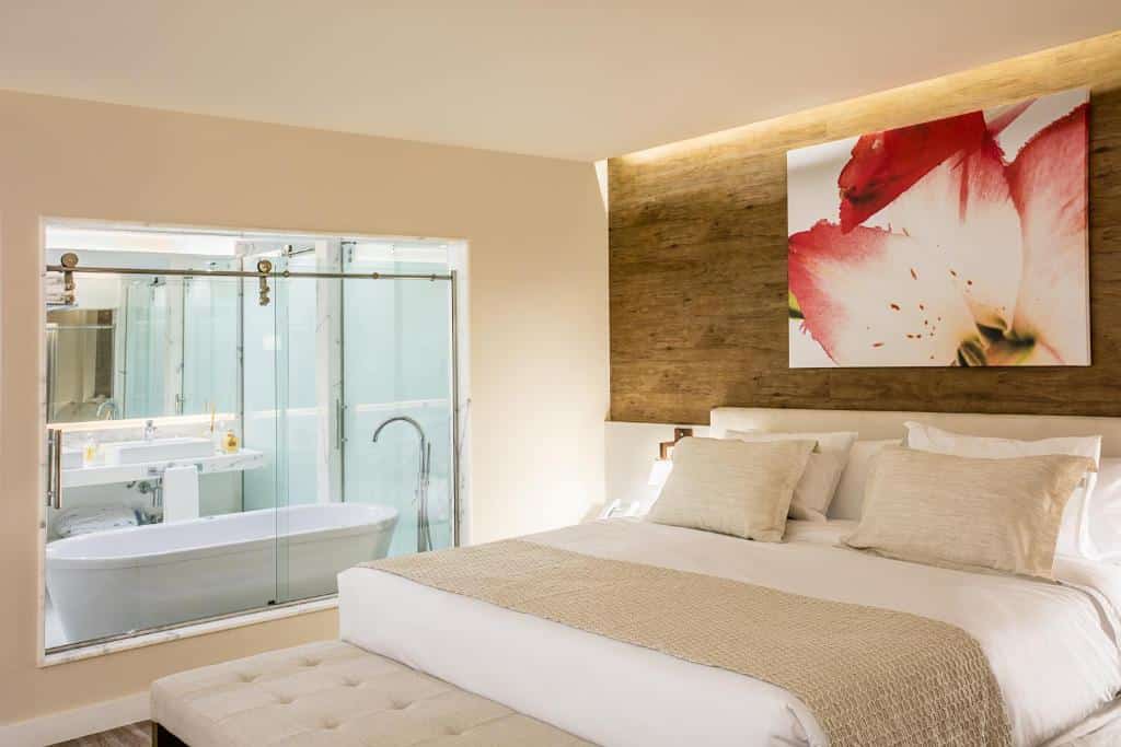 Quarto moderno no Vogue Square Fashion Hotel by Lenny Niemeyer com uma cama king-size, decoração bege, banheiro com paredes de vidro e uma pintura de flores na parede, para representar hotéis na Barra da Tijuca no RJ.