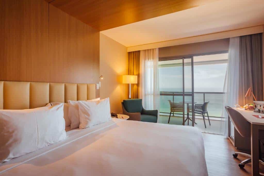 Quarto moderno do Laghetto Stilo Barra com uma cama grande, escrivaninha, poltrona, luminária de chão e varanda com vista para o oceano, para representar hotéis na Barra da Tijuca no RJ.