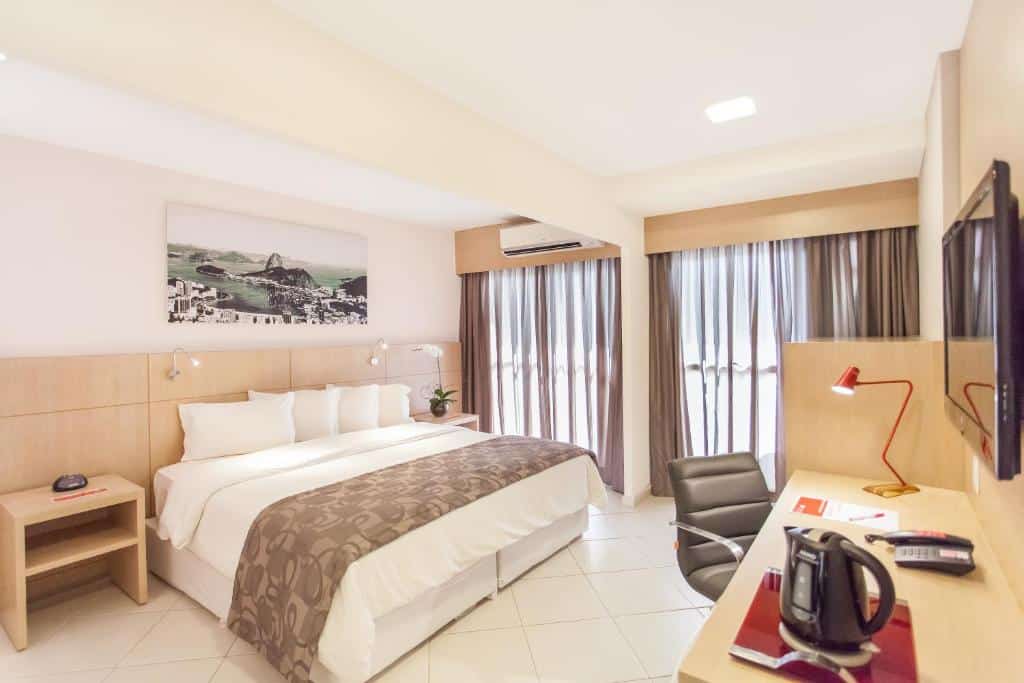 Quarto do Quality Barra da Tijuca com uma cama grande, escrivaninha, cadeira, TV, mesa de cabeceira e janelas com cortinas que permitem a entrada de luz natural, para representar hotéis na Barra da Tijuca no RJ.
