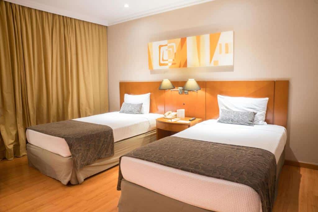 Quarto no Plaza Barra First com duas camas de solteiro, paredes bege, cortinas douradas, arte na parede, luminárias e uma mesa de cabeceira com telefone, para representar hotéis na Barra da Tijuca no RJ.