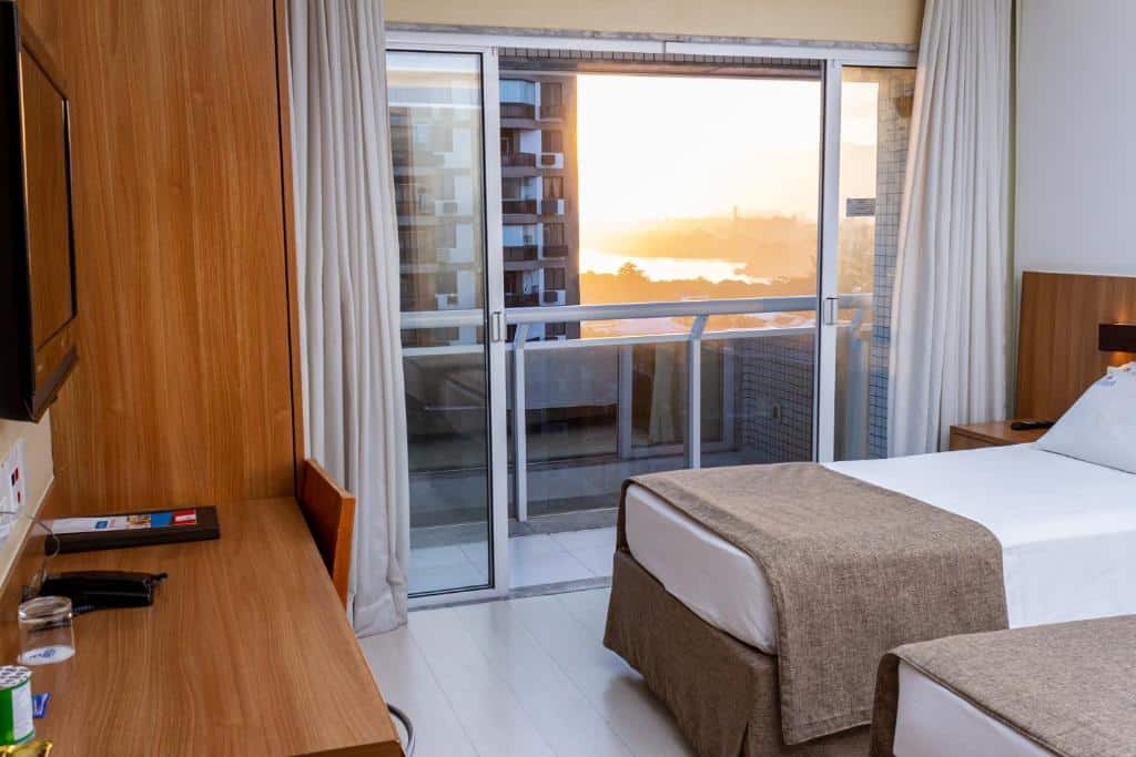 Quarto do Riale Brisa Barra com camas de solteiro, escrivaninha, TV e portas de vidro deslizantes que se abrem para uma varanda com vista para o pôr do sol, para representar hotéis na Barra da Tijuca no RJ.