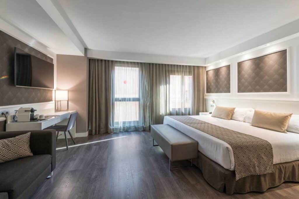 Quarto do Catalonia Magdalenes, uma das recomendações de hotéis no centro de Barcelona. A cama tem lençol e travesseiros bege e brancos com um banquinho bege aos pés. Há luminárias nas mesinhas de cabeceira dos dois lados da cama.
Na parede em frente há uma mesa de trabalho com cadeira, abajur e cafeteira. Há uma televisão na parede acima e uma potrona marrom ao lado.
Ao fundo do quarto, uma janela tem cortinas bege e brancas.