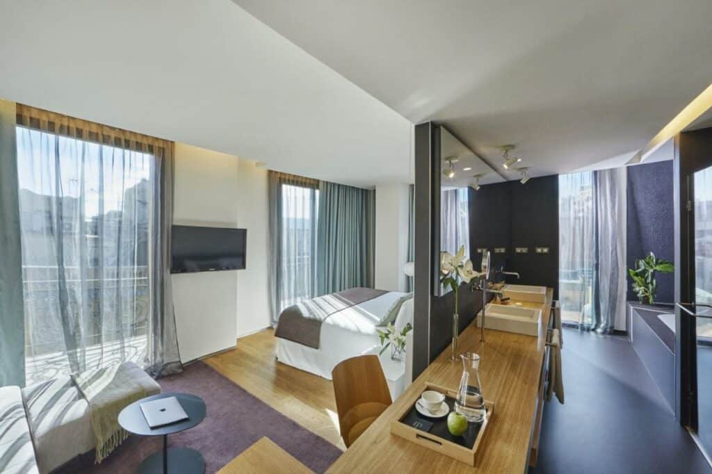 Quarto do Ohla Barcelona, uma das recomendações de hotéis no centro de Barcelona. Do lado esquerdo da imagem há uma cama de casal de frente para uma parede com duas janelas cobertas por cortinas cinzas transparentes. Uma televisão está de frente para a cama. Ao lado da janela no canto inferior esquerdo da foto há um banquinho com uma mesa em frente.
Uma parede separa o quarto do banheiro, e pode-se ver um espelho na parede acima das duas pias. Uma banheira está no fundo da imagem, e há outra janela no banheiro. Uma mesa de trabalho está ao ao lado da parede que separa os cômodos, e há uma bandeja com xícara, maça e uma garrafa d'água em cima.