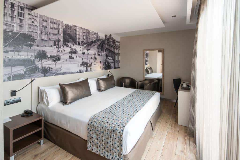 Quarto do Catalonia Born, uma das recomendações de hotéis no centro de Barcelona. Uma ampla cama de casal com mesinhas de cabeceira e luminárias dos dois lados encara uma janela coberta por cortinas brancas. Uma mesa de trabalho com cadeira e amenidades de café está ao lado da janela. Ao fundo do quarto há duas poltronas de couro perto de um espelho.