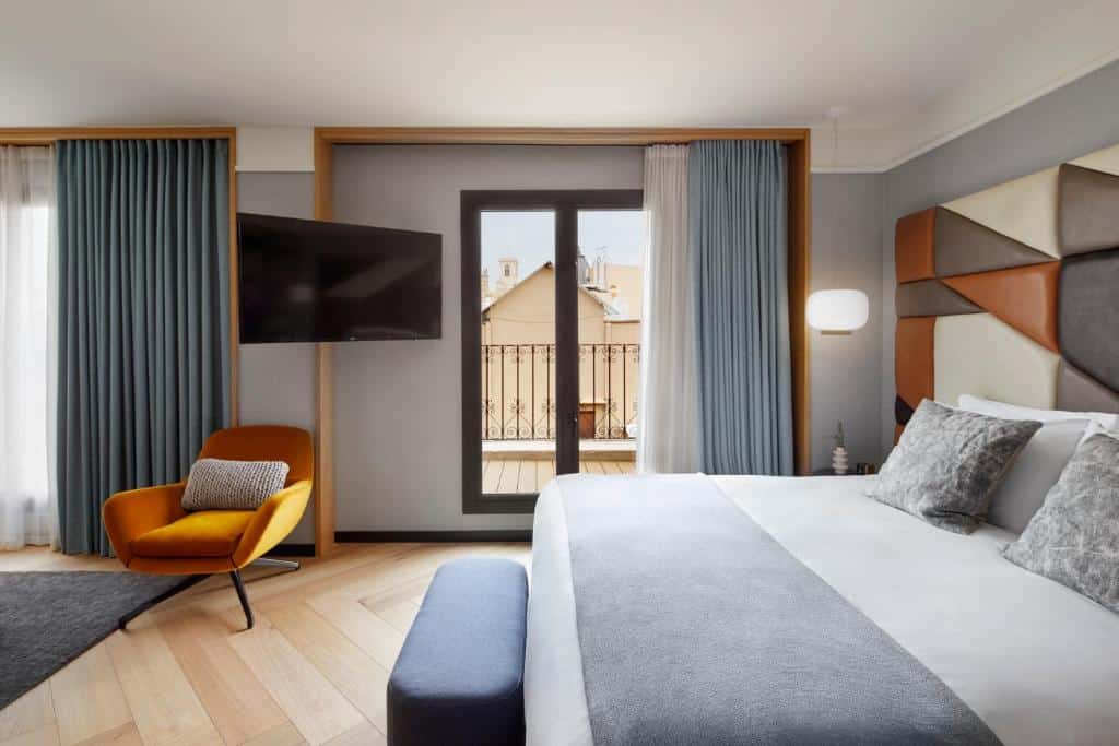 Quarto do Kimpton Vividora Hotel, uma das recomendações de hotéis no centro de Barcelona. A cama de casal está encostada em um painel colorido na parede direita, e tem um banquinho aos seus pés e mesinhas de cabeceira dos dois lados com luminária. Uma janela está no centro da parede com cortinas azuis e brancas abertas, e uma televisão encara a cama. Uma poltrona amarela está logo abaixo da TV, com um tapete cinza aos pés.