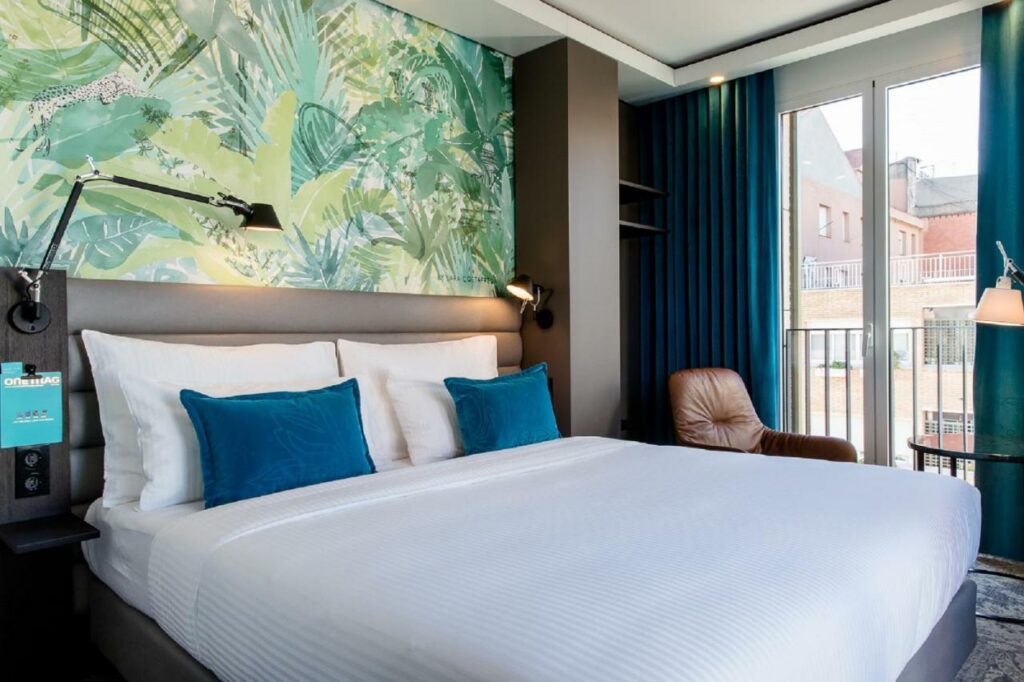 Quarto do Motel One Barcelona-Ciutadella, uma das recomendações de hotéis no centro de Barcelona. A cama está encostada em uma parede estampada com plantas e tem travesseiros brancos e azuis em cima. Há mesinhas de cabeceira com luminárias dos dois lados da cama, e na parede lateral uma janela está com as cortinas azuis abertas. Há uma poltrona de couro com mesinha e abajur ao lado da janela.