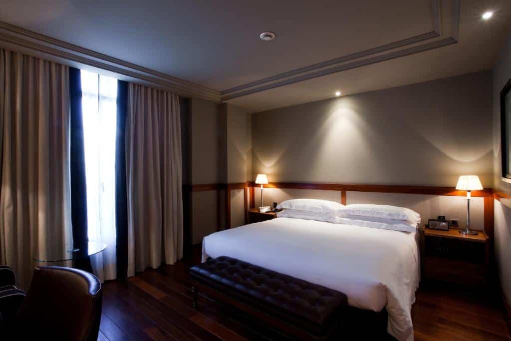 Quarto do Hotel 1898, uma das recomendações de hotéis no centro de Barcelona. A cama de lençol e travesseiros brancos tem abajures nas mesinhas de cabeceira dos dois lados, e há um banco de couro marrom aos seus pés.
Há uma mesa de trabalho com cadeira de frente para a cama, e ao lado uma janela fechada por cortinas beges.
