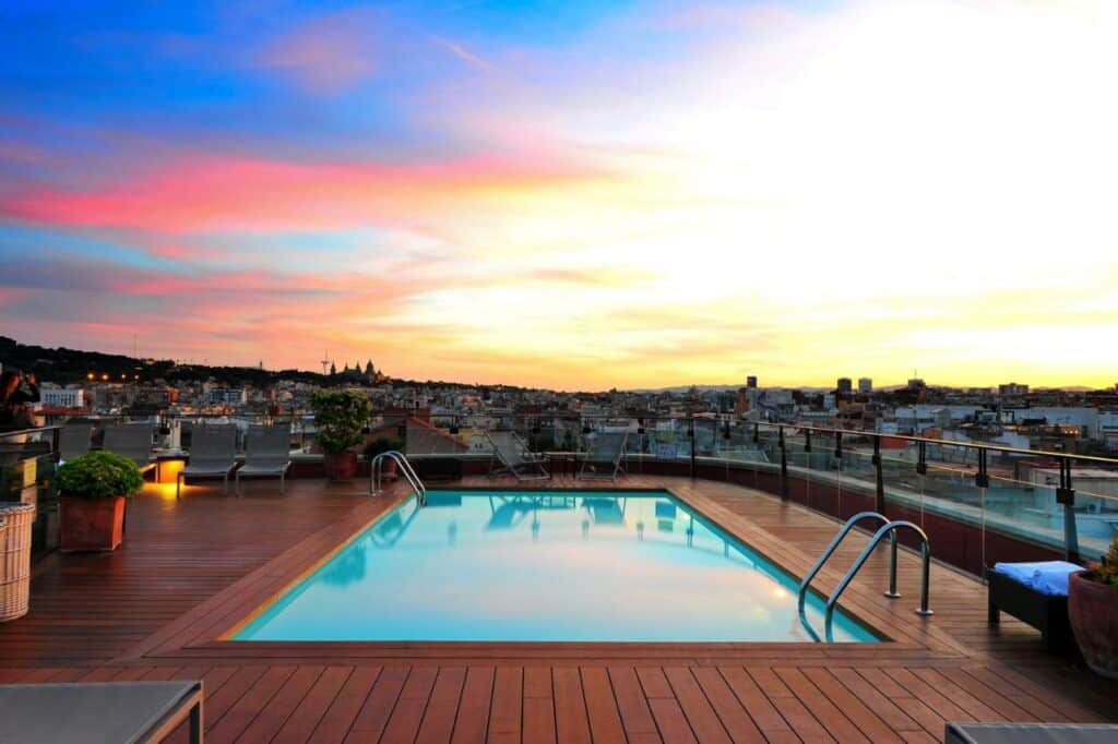 Piscina do Hotel 1898. Há um deque de madeira por toda a cobertura de onde é possível ver a cidade. Há algumas plantas e espreguiçadeiras ao redor, e duas escadas levam à piscina. Nuvens cor de rosa se mesclam ao azul do céu.