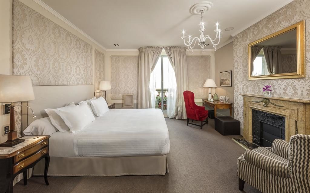 Quarto do Hotel El Palace Barcelona, uma das recomendações de hotéis no centro de Barcelona. A cama com lençol e travesseiros brancos está encostada na parede esquerda, e há abajures nas mesinhas de cabeceira dos dois lados da cama. Há uma lareira na parede em frente com um espelho logo acima. De um lado há uma poltrona listrada, e do outro uma mesa de trabalho com poltrona vermelha, abajur e amenidades de chá está próxima da janela que tem cortinas bege. Um lindo lustre está pendurado no centro do quarto.