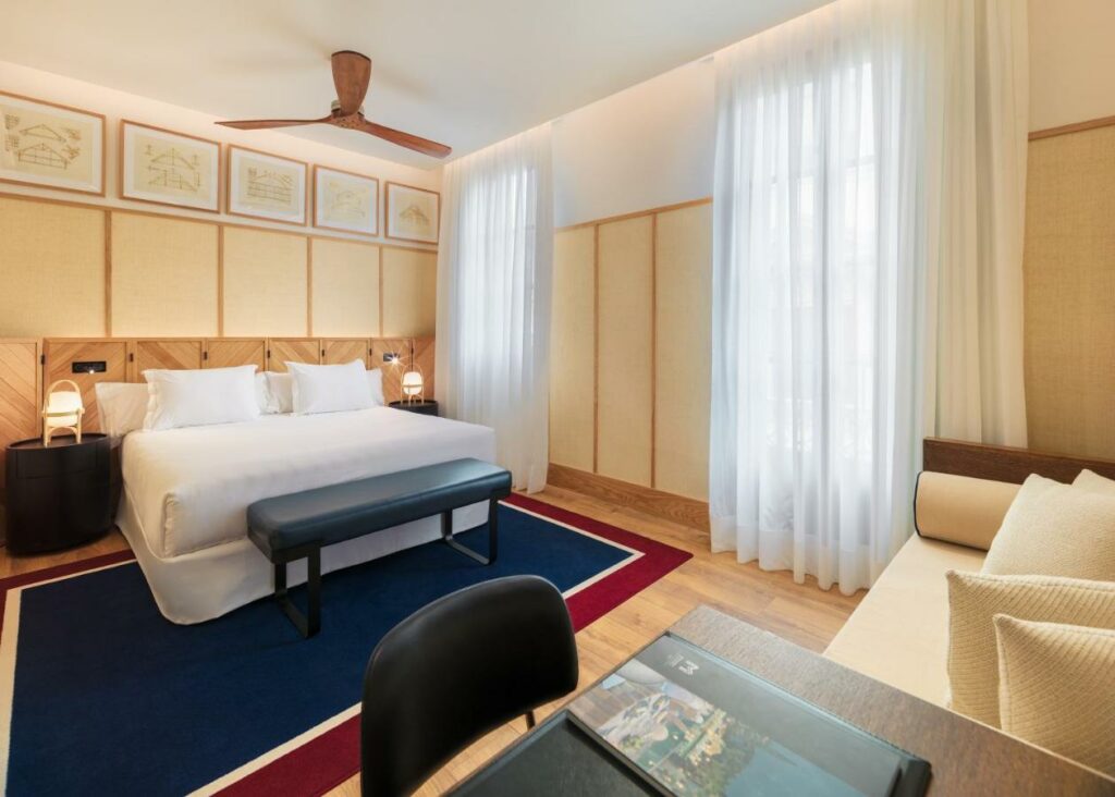 Quarto do H10 Madison, uma das recomendações de hotéis no centro de Barcelona. Uma cama de casal tem mesinhas de cabeceira com abajur dos dois lados, e um banco aos seus pés. Há um ventilados de teto em cima da cama, e um tapete azul marinho e vermelho embaixo dela. De frente há um sofá e uma mesa de trabalho com cadeira. Na parede ao lado há duas janelas cobertas por cortinas brancas diáfanas.