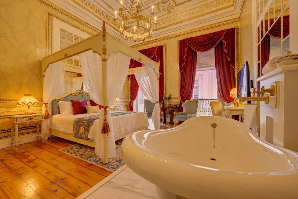 Quarto luxuoso do Palácio das Especiarias com duas sacadas com cortinas vermelhas, o ambiente inteiro remete a realeza, há um cama de casal com um bangalô com tecidos brancos, há um lustre dourado e também uma banheira de hidromassagem, para representar hotéis no centro de Lisboa.