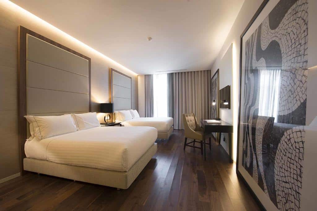 Quarto do BessaHotel Liberdade com duas camas de casal, uma janela com cortinas cinzas, uma mesa de escritório com uma cadeira estofada e, de frente para as camas, há uma televisão, além de dois grandes quadros, para representar hotéis no centro de Lisboa.