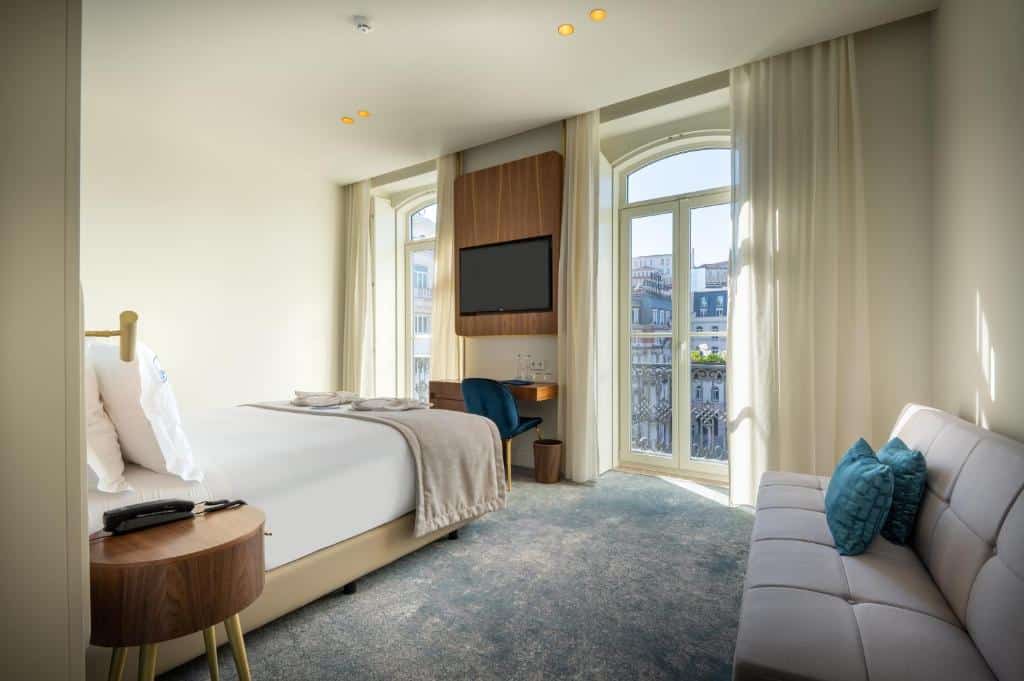 Quarto do Blue Liberdade Hotel com duas sacadas e, entre elas, há uma televisão e uma mesa de escritório com uma cadeira, no centro do quarto está uma cama de casal e do lado direito há um pequeno sofá com duas almofadas.