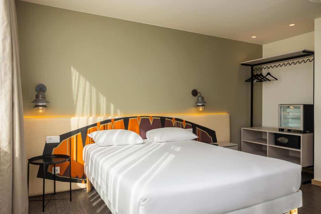 Quarto do Hotel Ibis Styles Lisboa Liberdade com uma cama de casal, um pequeno frigobar junto de um armário de conceito aberto com cabideiro.