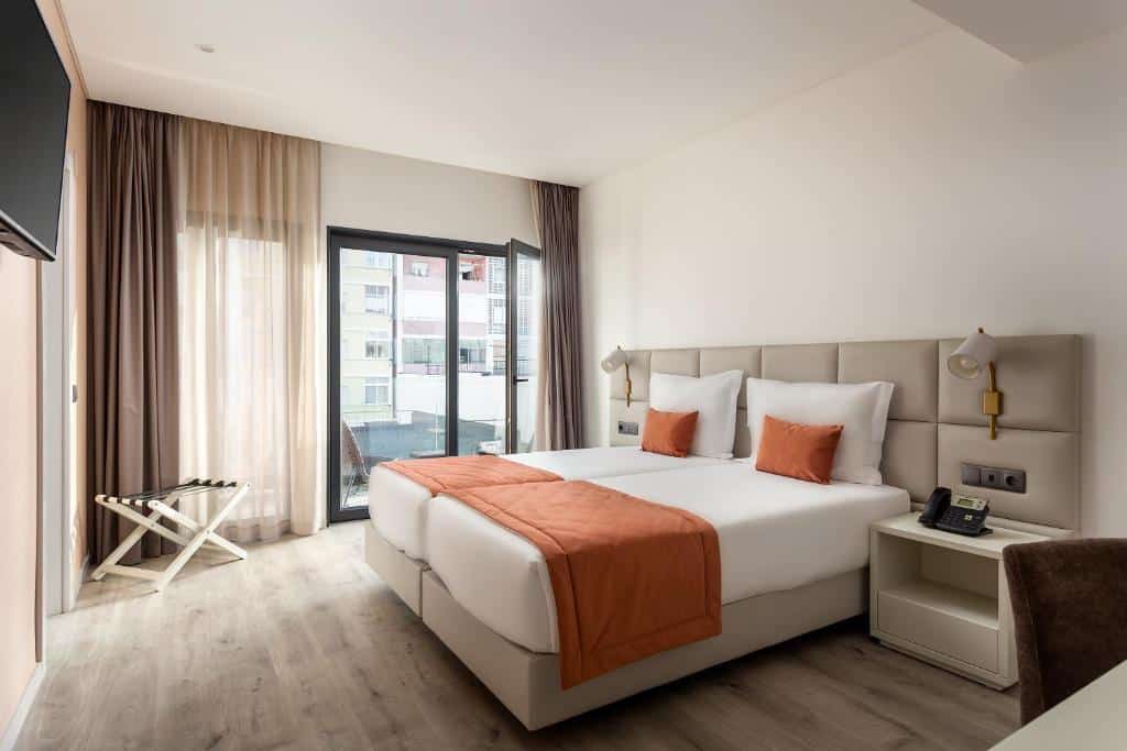 Quarto do Empire Marquês Hotel com uma pequena sacada com duas cadeiras, há duas camas de solteiro com uma cabeceira estofada com luminárias presas, há duas mesinhas de cabeceira também, e de frente para a cama há uma televisão, para representar hotéis no centro de Lisboa.