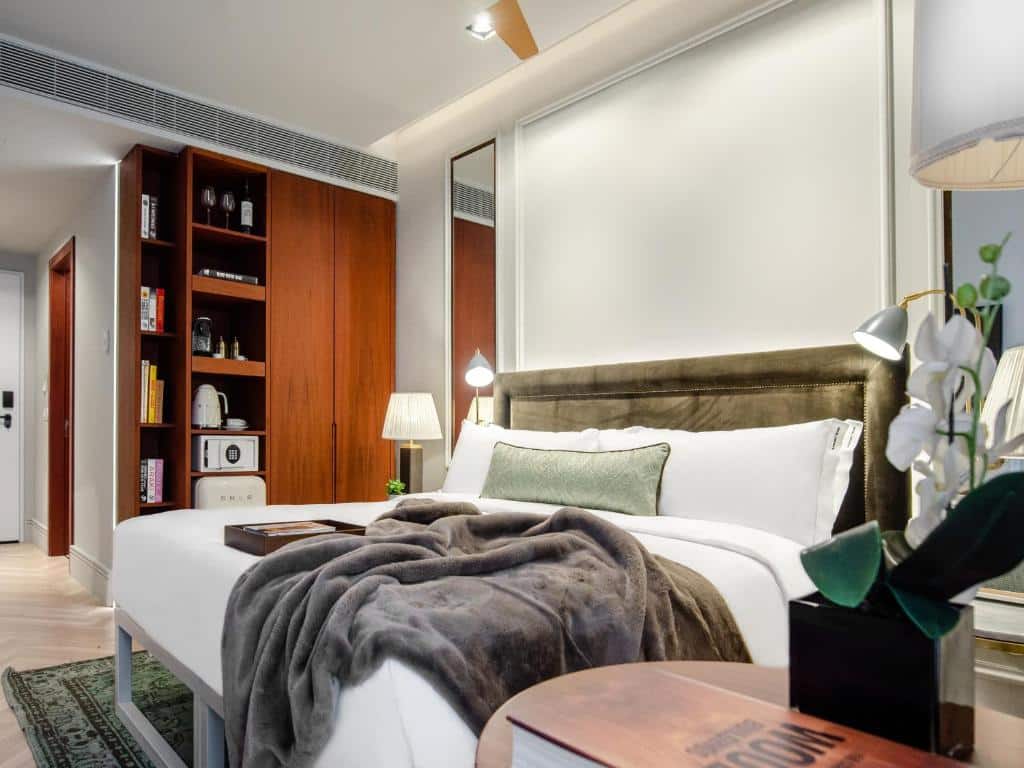 Quarto do Browns Avenue Hotel com uma cama de casal com almofadas e um cobertor, do lado esquerdo há uma armário pequeno com duas portas e uma prateleira com livros, um frigobar, comodidades para preparar café e um cofre, há também luminárias nas laterais da cama.