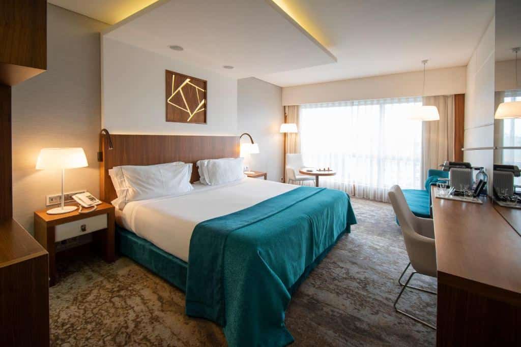 Quarto do EPIC SANA Lisboa Hotel com uma cama de casal, uma sacada com cortinas, de frente para a cama há uma mesa de escritório e uma cadeira estofada, nas laterais da cama há mesinhas de cabeceira com abajures.