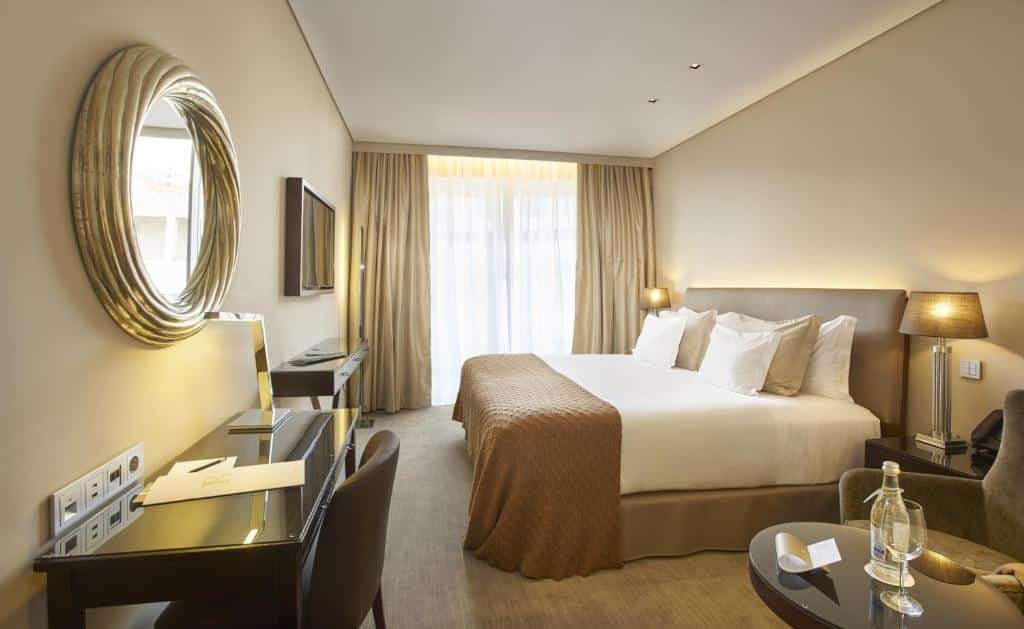 Quarto do PortoBay Liberdade com uma cama de casal, uma sacada com cortinas, na parede de frente com a cama há um pequeno móvel e uma televisão acima, assim como há uma pequena mesa com uma cadeira estofada e uma mesa de escritório, para representar hotéis no centro de Lisboa.