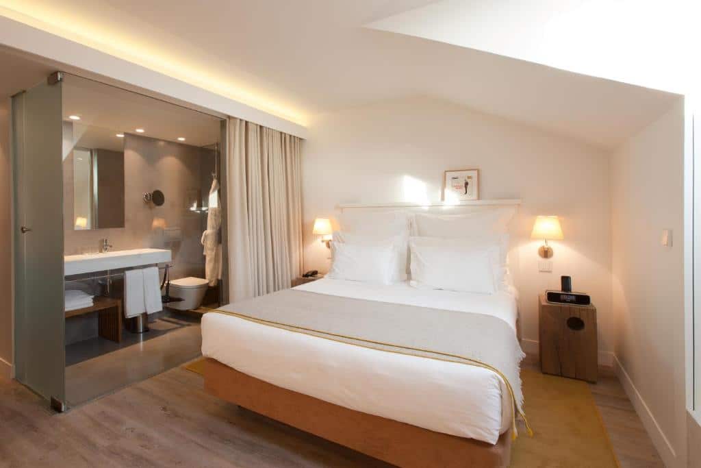 Quarto do Memmo Alfama – Design Hotels com uma cama de casal, logo ao lado, separado por um cortina, há uma banheiro com pia, vaso, roupões e espelho, do lado direito do cômoda há uma janela.