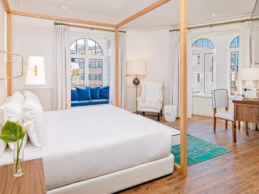 Quarto chique do H10 Duque de Loule com uma cama de casal ampla, duas janelas, sendo que numa delas há um pequeno sofá com almofadas, há também uma poltrona branca com um abajur de chão, para representar hotéis no centro de Lisboa.