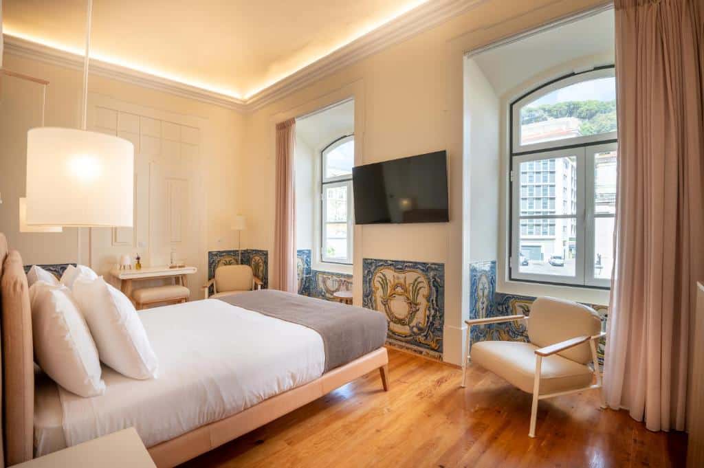 Quarto do Memoria Lisboa FLH Hotels com uma cama de casal de frente para uma parede com duas janelas e, entre elas, há uma televisão presa, do lado esquerdo do quarto há uma pequena bancada e uma cadeira estofada, o chão imita madeira e algumas paredes tem azulejos coloridos.