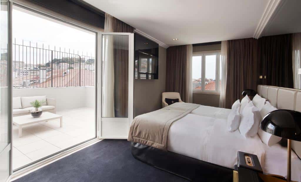 Quarto do Altis Avenida Hotel com uma varanda com um sofá e uma mesinha, do lado de dentro há uma cama de casal com travesseiros, do lado esquerdo há uma janela com cortinas e uma poltrona, de frente para a cama há uma televisão na parede, para representar hotéis no centro de Lisboa.