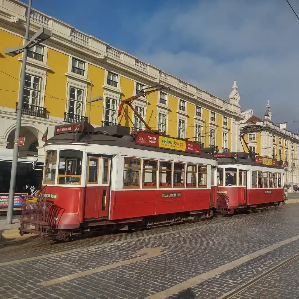 Um bonde vermelho antigo está estacionado em trilhos de paralelepípedos em frente a um prédio amarelo com acabamento branco, em Lisboa.