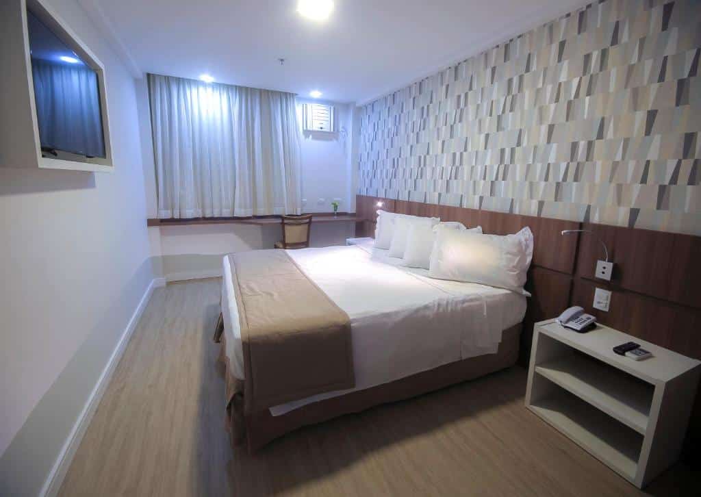 Um quarto de hotel moderno com uma cama de casal, uma parede com detalhes estampados, uma escrivaninha com cadeira e uma TV na parede. Este é um dos hotéis no centro do rio de janeiro.