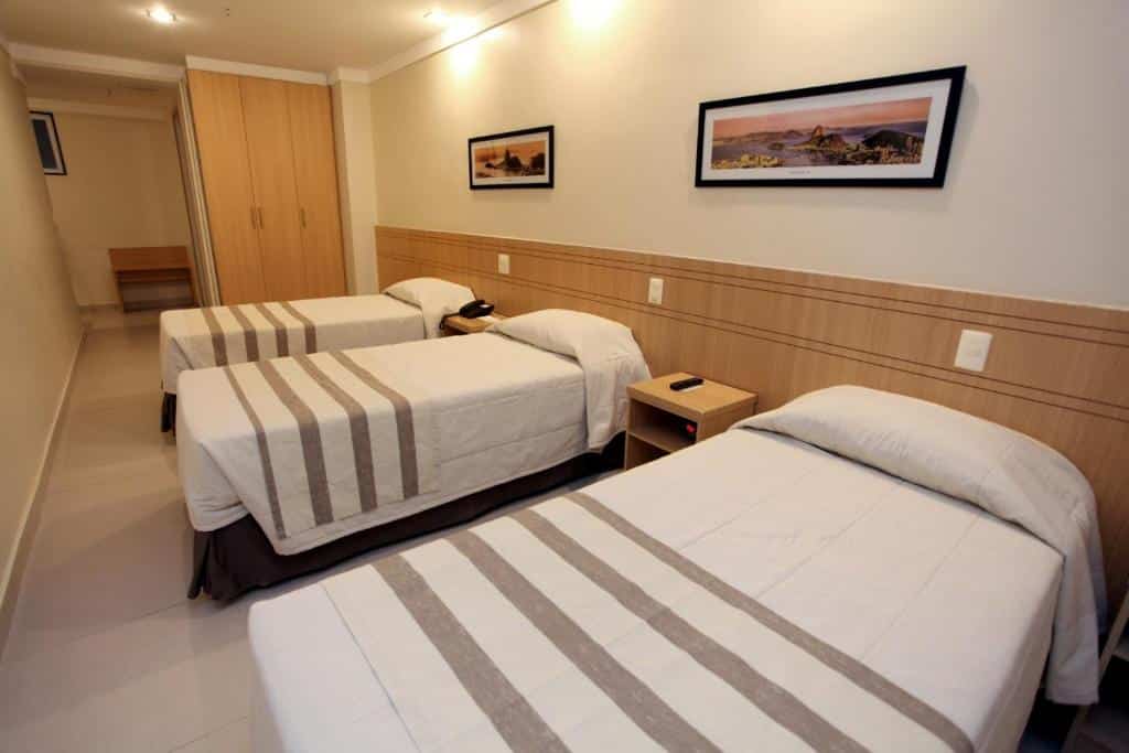 Um quarto de hotel com três camas de solteiro, roupa de cama listrada, móveis de madeira e dois quadros de paisagens na parede.